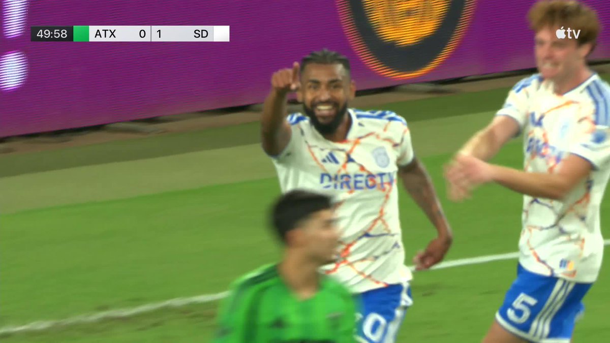 A San Diego FC player in a white "DIRECTV" jersey celebrates with a pointed finger gesture after scoring, with the scoreboard showing ATX 0-1 SD at 49:58, matching Aníbal Godoy's go-ahead goal for San Diego on the road. A teammate wearing #5 runs over to join the celebration as an Austin FC player in green walks past dejectedly.