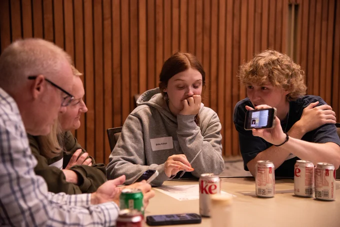 Four people gather around a table in a wood-paneled room; a young man shows his phone as others listen.
