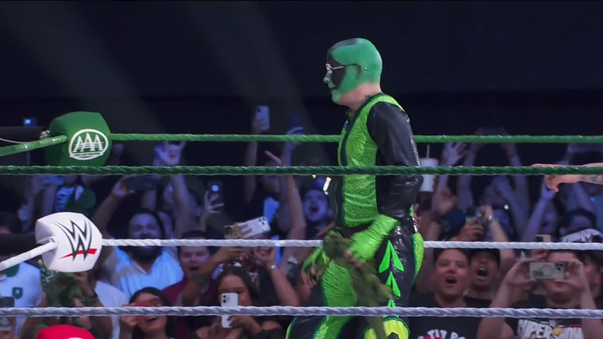 A luchador wearing a green mask and matching green-and-black costume, identified as "Mr. Iguana," stands in a wrestling ring surrounded by cheering fans holding up phones, with the AAA and WWE logos visible on the ring turnbuckles.