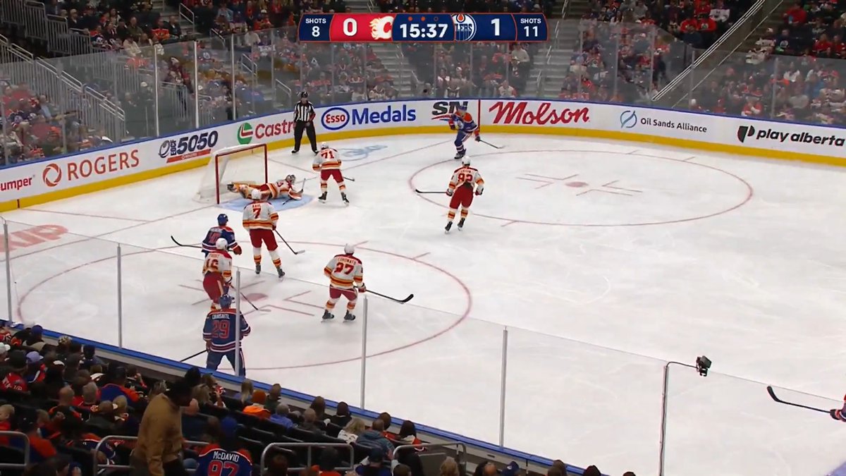 A hockey game is underway between the Flames (in white jerseys) and Oilers (in blue jerseys), with the scoreboard showing the Flames leading 1-0 with 15:37 remaining in the period, as the Oilers' goaltender sprawls across the crease to make a save while Flames players converge on the net. The caption "DENIED" aptly captures the goalie's impressive defensive stop, preventing a likely Flames scoring opportunity.
