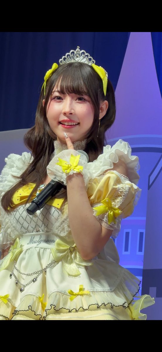 A young woman with a yellow bow in her hair and a tiara smiles while holding a microphone, wearing an elaborate yellow and white frilly dress with a 'Meltymoon' sash.