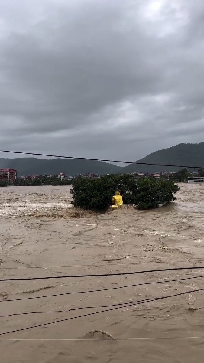 A yellow Buddha statue sits amid flooded, muddy brown waters flowing rapidly around trees and a distant town under a gray, cloudy sky.