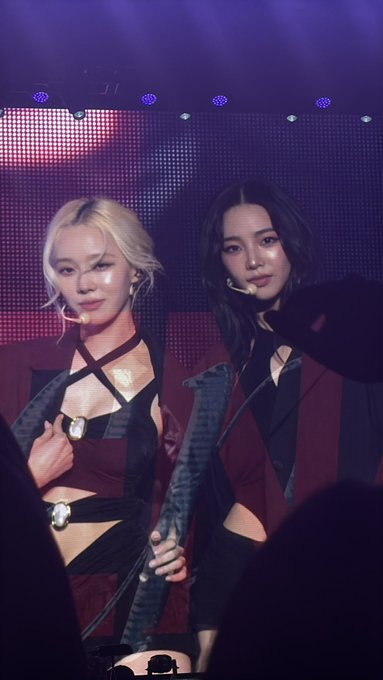 Two female K-pop performers, one blonde and one with dark hair, wear matching red and black outfits on a stage.