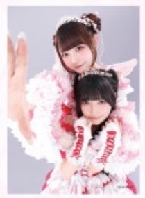 Two young women wearing matching pink and white frilly idol costumes pose closely together for a photo.