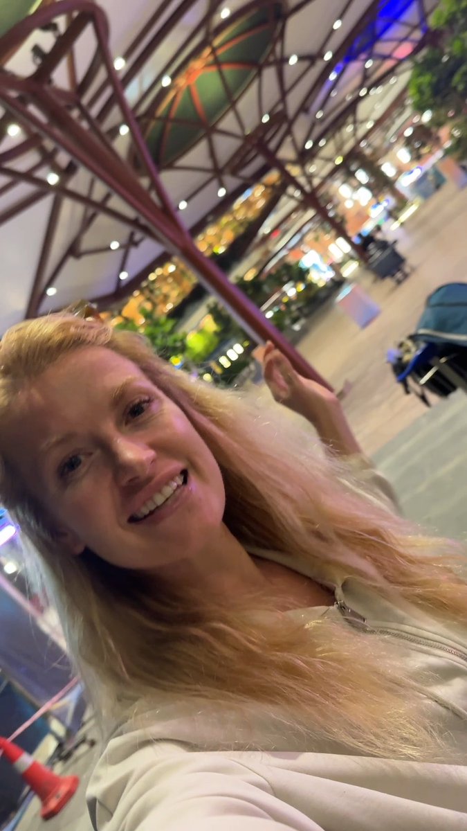 A smiling blonde woman takes a selfie in what appears to be a shopping mall, with hanging decorations and storefronts visible in the background.