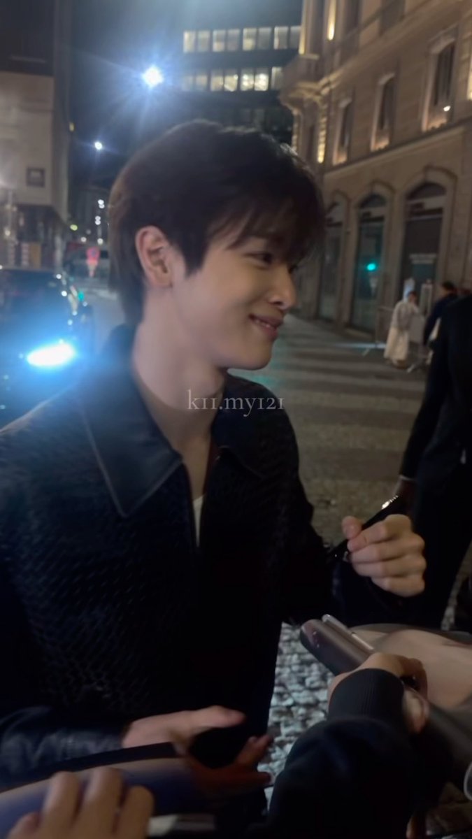 A young man smiles warmly while walking down a nighttime city street, seemingly signing autographs for fans. The watermark "kil.my121" appears on his jacket, matching the post's playful warning that his laughter is too obviously endearing.