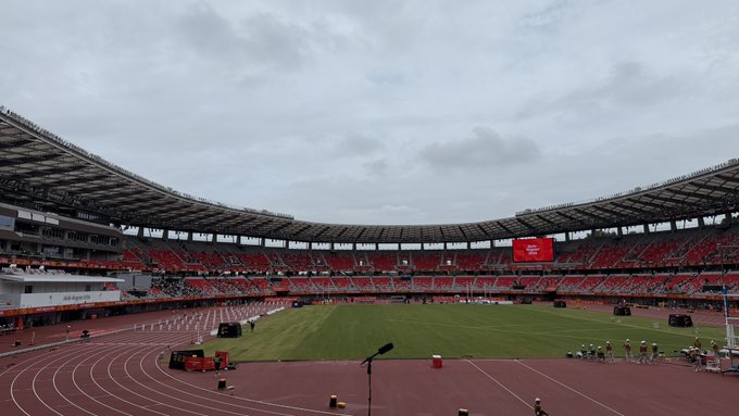 A wide view of a large stadium with red seats, a running track, and a green field, under an overcast sky with some equipment set up on the field.