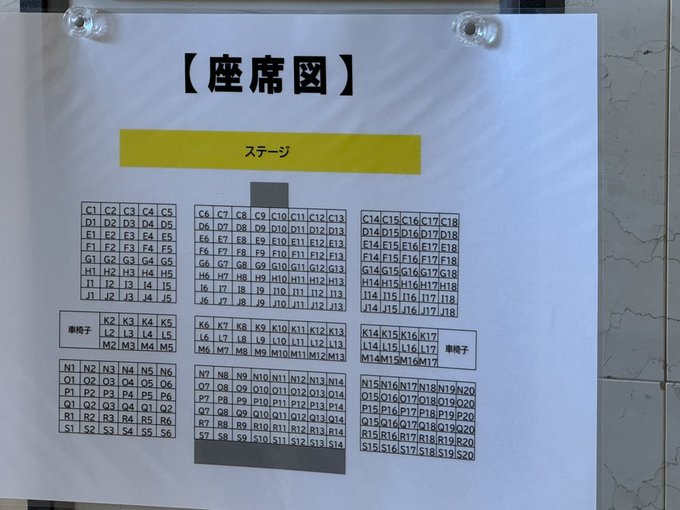 A Japanese seating chart posted on a wall, with '【座席図】' (Seating Chart) heading and a yellow stage indicator showing rows of seats labeled with codes.