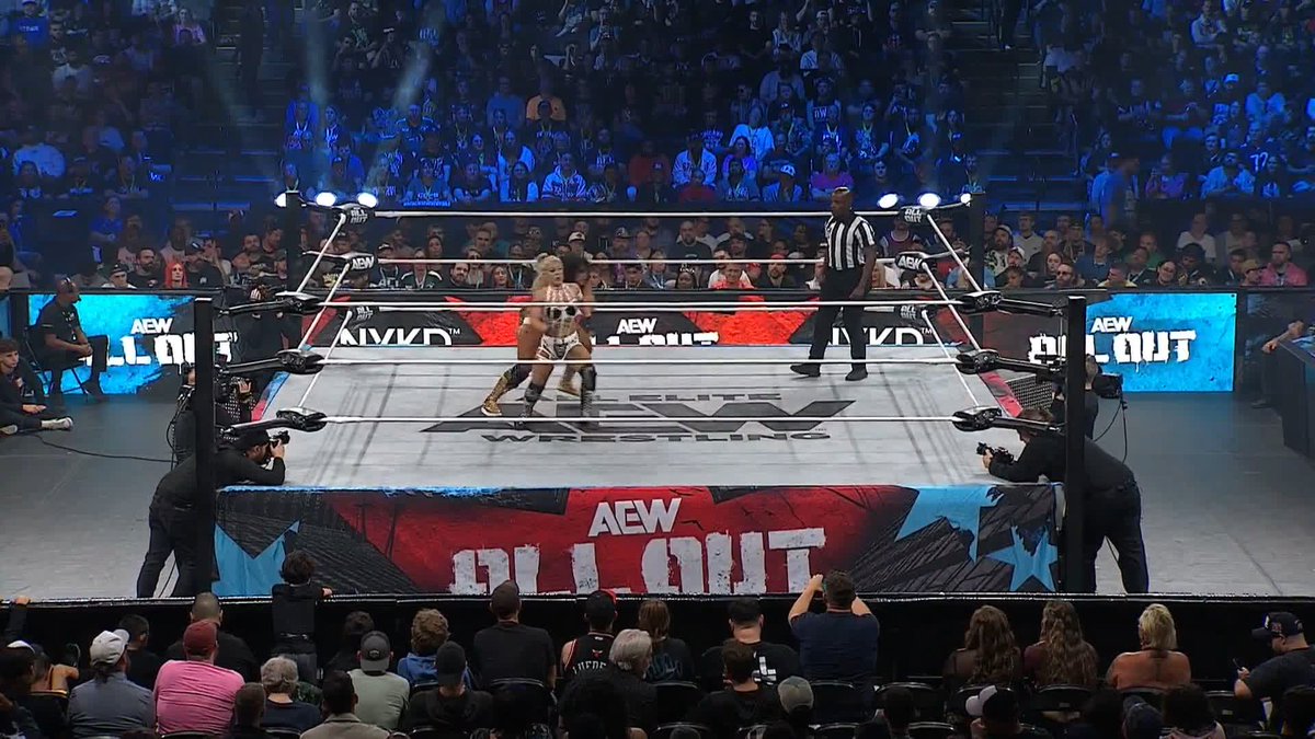 Two professional wrestlers face off in an AEW wrestling ring during an All Out event, with a referee and cameramen surrounding them as a large crowd watches.