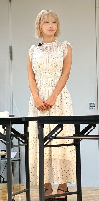 A young woman with short blonde hair stands indoors behind a black metal railing, wearing a white sleeveless dress with small dark polka dots.