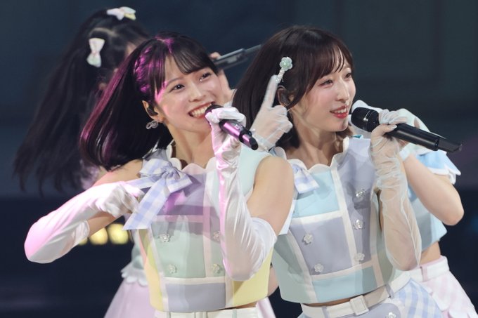 Three female performers in pastel-colored idol costumes with white gloves smile and sing into microphones during a live concert performance.