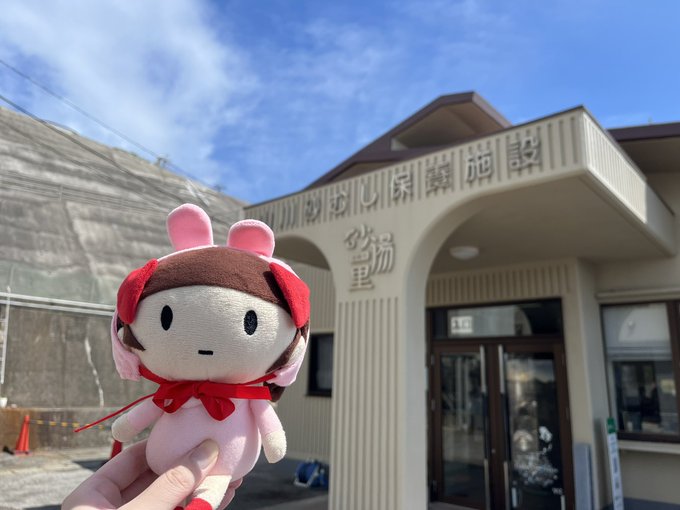 A hand holds up a pink stuffed mascot toy wearing a red hood in front of a building with Japanese signage including '砂湯' (sand bath) and '心保養施設' (heart recuperation facility).