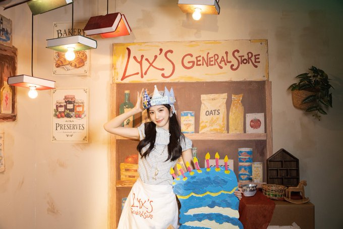 A young woman wearing a paper crown and "LYX'S General Store" apron poses next to a decorated blue birthday cake in a themed bakery backdrop with painted signs reading "BAKERY," "PRESERVES," and "LYX'S GENERAL STORE."