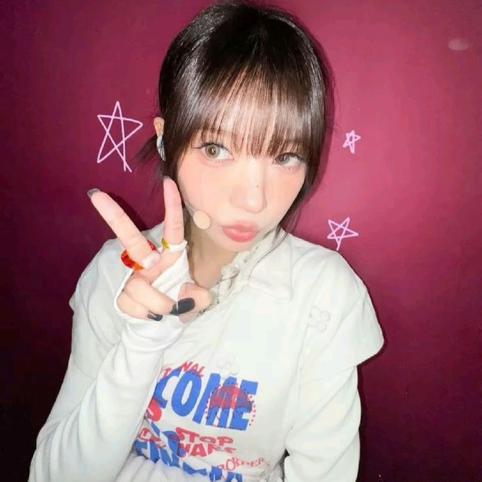 A young woman with short dark hair makes a peace sign, wearing a white Star Wars shirt against a purple background with drawn stars.