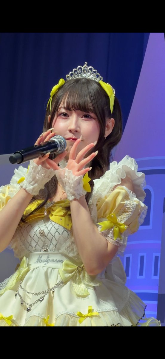 A young woman wearing an elaborate yellow and white frilly idol costume with a tiara holds a microphone while performing on stage.