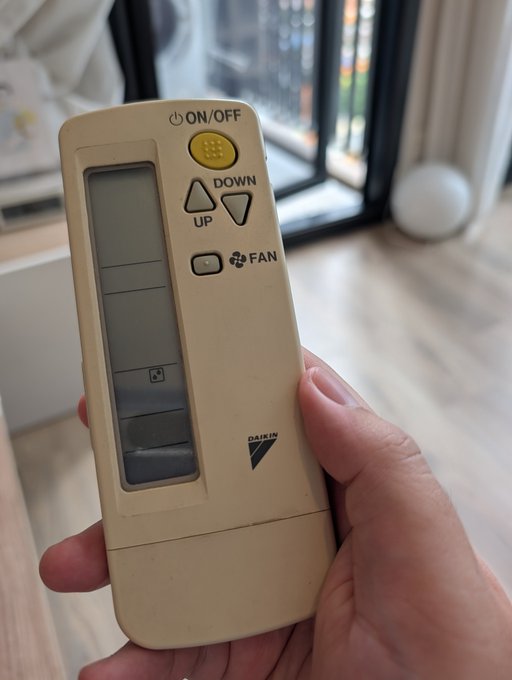A hand holds a yellowed Daikin air conditioner remote control featuring ON/OFF, DOWN/UP, and FAN buttons, corresponding to the post about running the AC in Dry mode frequently to reduce humidity and eliminate the sticky feeling in the room.