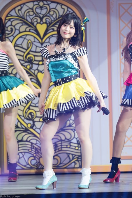 A female performer in a colorful yellow, teal, and black-and-white striped idol outfit poses on stage, with a watermark '@photo_nyonyo' visible.