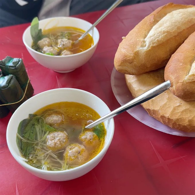 Two bowls of Vietnamese meatball soup with broth, herbs, and noodles are served alongside a plate of crusty baguette rolls on a red table.