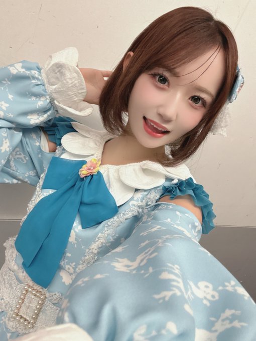 A young woman smiles while taking a selfie in a blue and white frilly outfit with a large blue bow and lace details, styled in an anime-inspired fashion.
