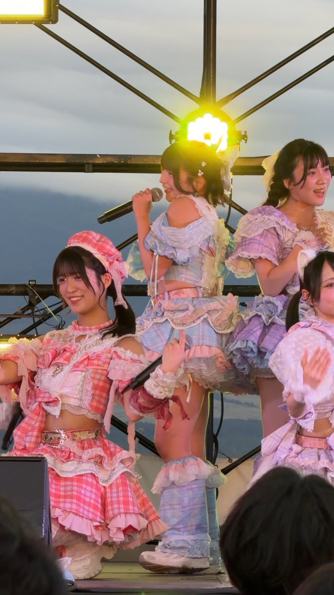 Four young performers in elaborate pastel-colored frilly idol costumes perform on an outdoor stage at SPARK 2026 in Yamanakako, with the central figure in a blue lace outfit holding a microphone during what the caption identifies as the "Koropokkur Sisters' Drop Pre-Chorus." The other members wear coordinated pink and purple checkered outfits, smiling and dancing under stage lights against a cloudy sky backdrop.