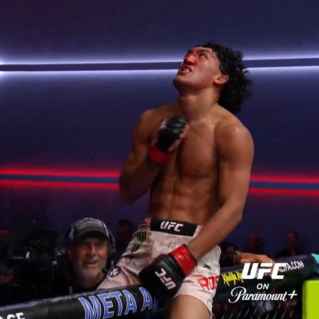 UFC fighter Raul Rosas Jr. celebrates dramatically with his head tilted back and glove raised in victory inside the octagon, wearing shorts branded "UFC" and "Monster," following his shocking win over Barcelos, who suddenly collapsed without being struck. The broadcast graphics show "UFC on Paramount+" along with "UFCVEGAS121" sponsorship banners, matching the post describing the eerie finish that left the MMA community concerned.