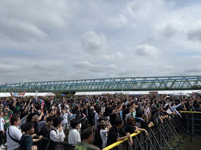 A large crowd of fans with raised hands gathers at an outdoor music festival, with a green bridge visible in the background under a cloudy sky.