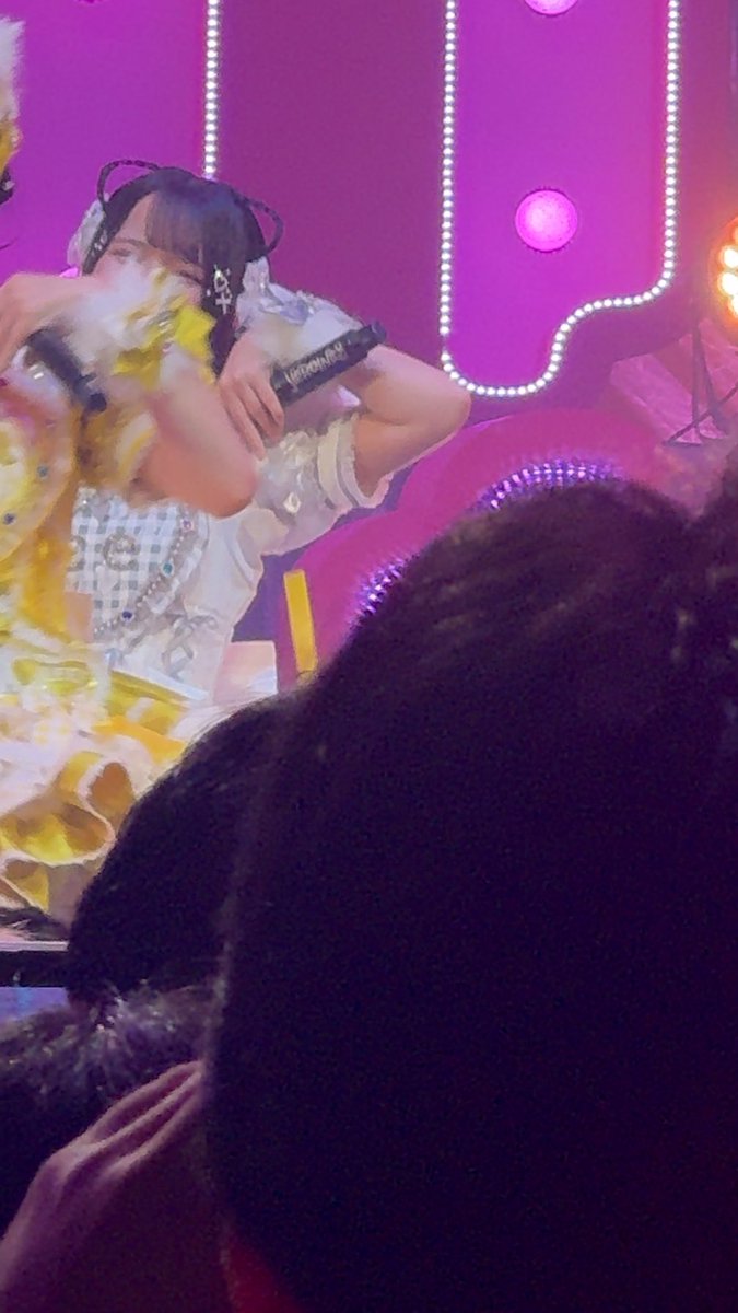 A performer in a yellow and white frilly outfit with cat-ear headphones plays a guitar on a brightly lit stage with pink LED lights, while an audience member watches from the foreground.