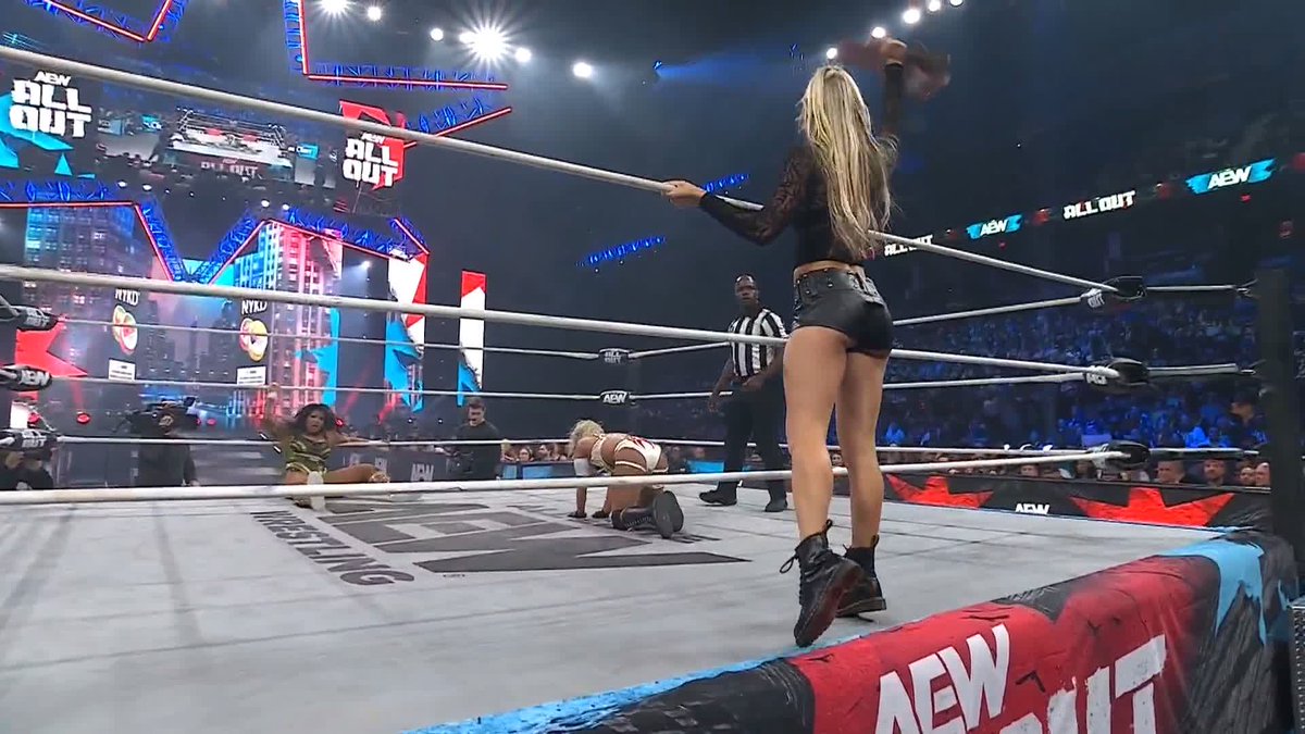 A scene from an AEW wrestling event shows a blonde woman in black shorts standing on the ring apron holding an object above her head, while a referee and other wrestlers are visible inside the ring under bright 'ALL OUT' signage.
