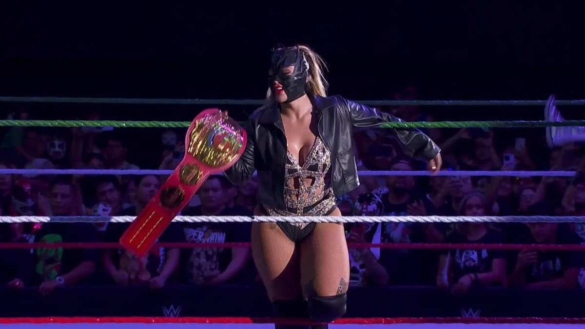 A wrestler wearing a black mask, leather jacket, and embellished bodysuit holds up a red and gold championship belt inside a wrestling ring, with fans visible in the background. The caption connects to the post by showcasing one of the competitors in the AAA three-way match on Fox.