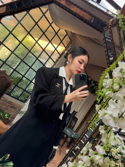 A stylish woman in a black blazer with a Chanel brooch and a white-collared shirt takes a mirror selfie with her phone in an elegant indoor setting with white floral arrangements.