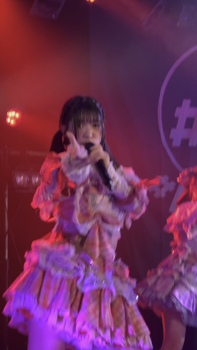 An idol performer in a ruffled pink and white frilly dress sings into a microphone on a stage lit with red lighting, making a pointing gesture toward the camera.