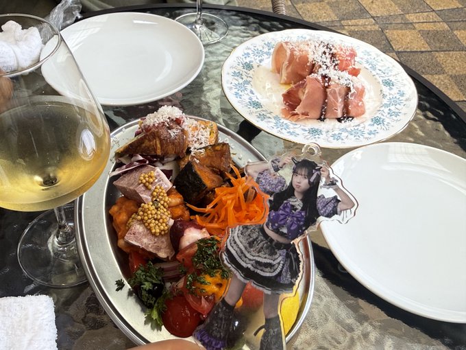 A dining table features a plate of assorted foods including prosciutto, vegetables, and meat, with a clear acrylic standee of an idol in a gothic dress placed among the dishes.