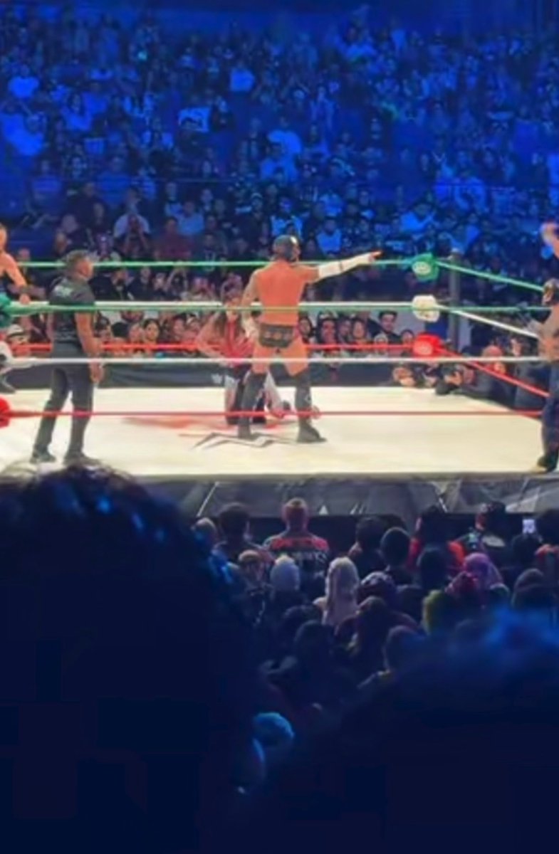 A masked wrestler, resembling CM Punk, points toward the crowd inside a brightly lit lucha libre ring with red, green, and white ropes, surrounded by a packed arena bathed in blue light. The scene captures the theatrical energy of the match referenced in the "#WoeldsCollide" post, with another wrestler seated behind him and spectators cheering from the stands.