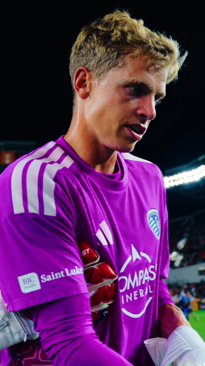 A young goalkeeper with blonde hair, wearing a purple Sporting Kansas City jersey with "Saint Luke's" and "Compass Mineral" sponsorships visible, looks down after a match. The image aligns with the caption "Stef gets his shutout," suggesting goalkeeper Stefan Cleveland earned a clean sheet victory.