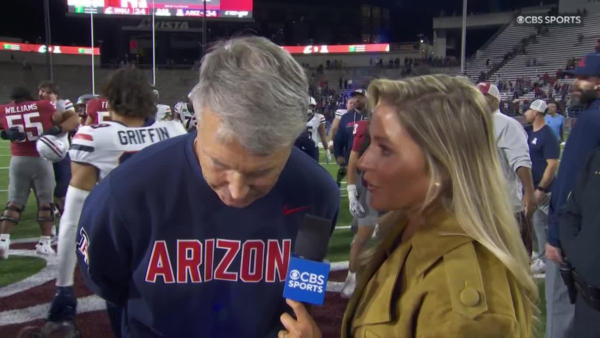 A CBS Sports reporter interviews Arizona head coach Brent Brennan, who wears a navy "ARIZONA" sweatshirt and looks down thoughtfully while holding a CBS Sports microphone on the field postgame, with the scoreboard showing ARIZ 34 and players like "WILLIAMS 55" and "GRIFFIN" visible in the background, capturing the moment Brennan praises his quarterback Noah Fifita.