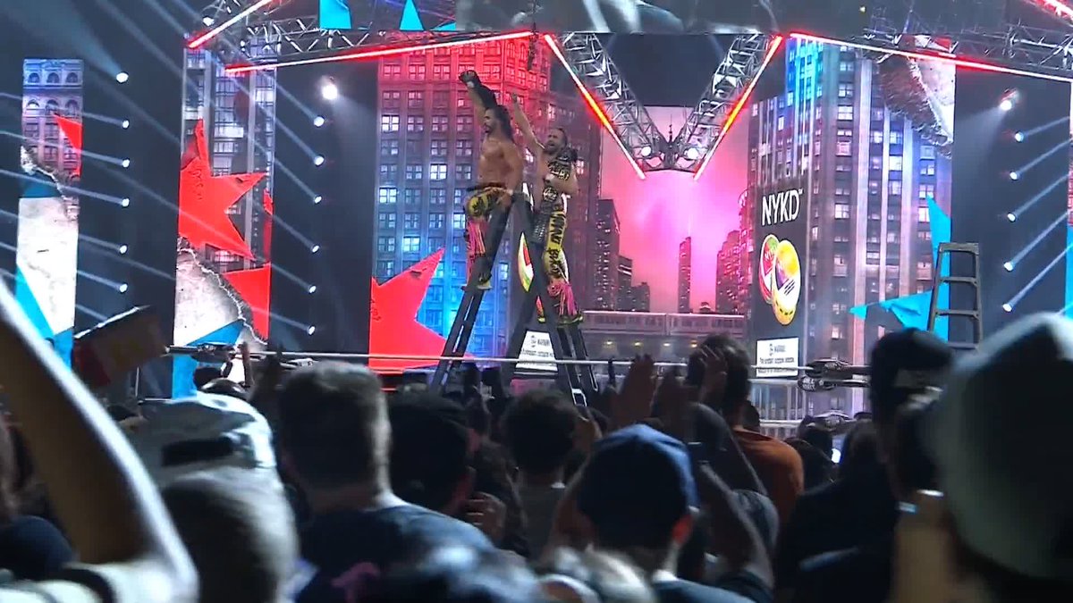 Two wrestlers stand atop a ladder in a wrestling ring, arms raised in victory as the crowd cheers below. The stage backdrop displays "NYKD" alongside a New York City skyline graphic, and the scene captures the Young Bucks celebrating their AEW World Tag Team Championship win after a Tables, Ladders, and Chairs match.