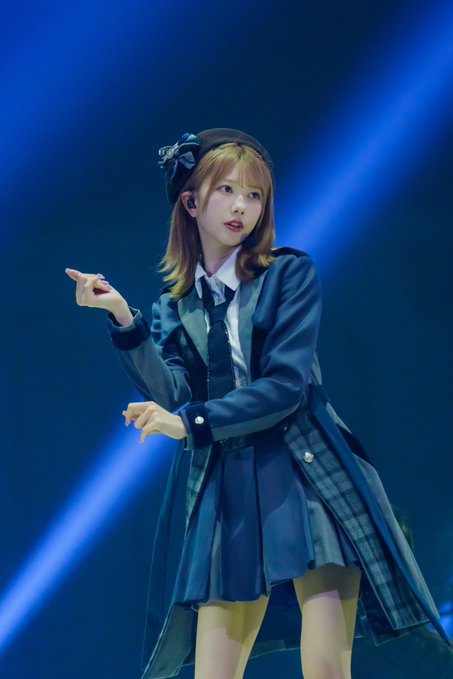 A young woman performs on stage in a dark school uniform-inspired outfit with a beret, pointing while singing under blue lighting.