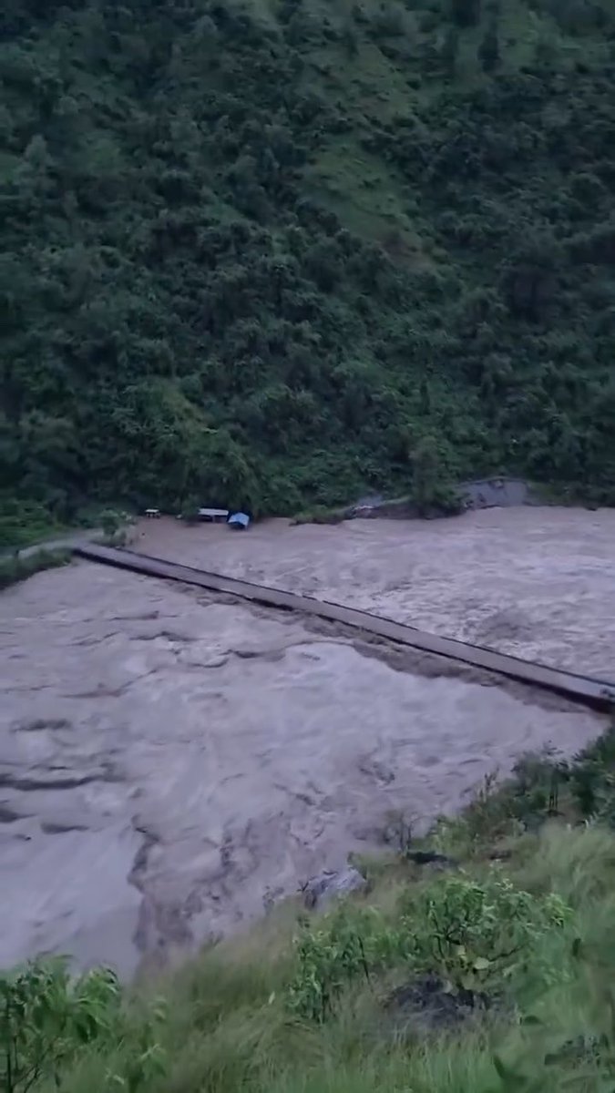 An aerial view of a flooded, muddy river rushing under a suspension bridge in a forested mountainous area.
