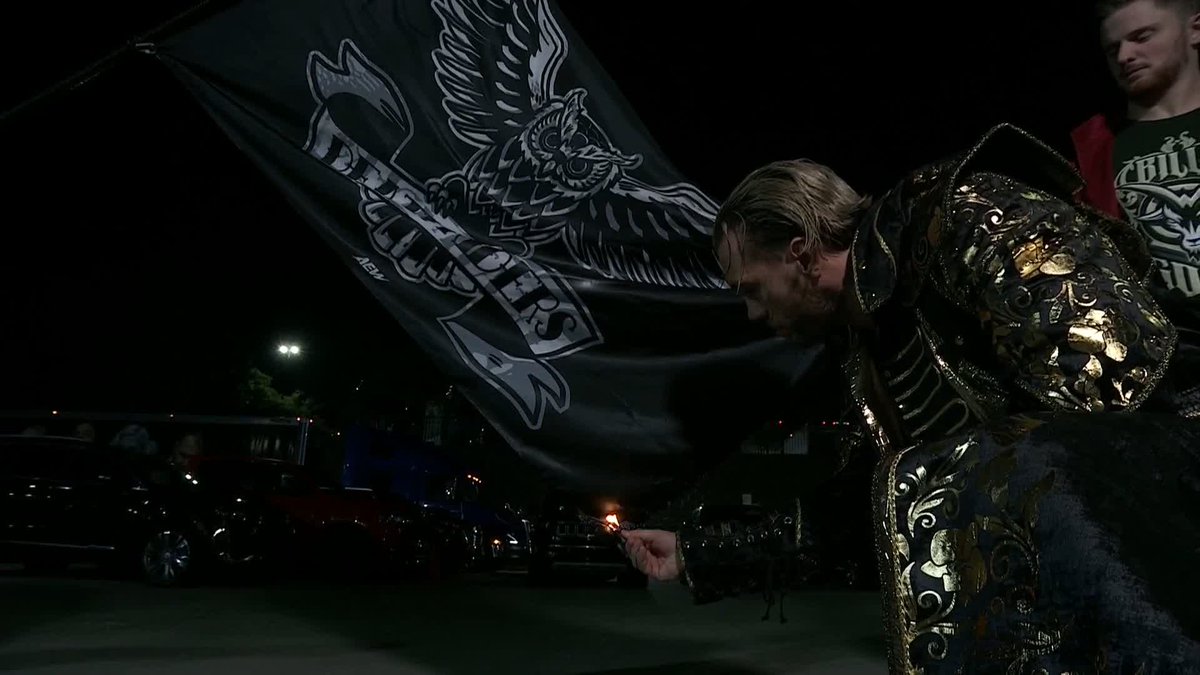 A wrestler in an ornate black and gold costume holds a large 'BULLET CLUB' flag at night, with vehicles visible in the background.