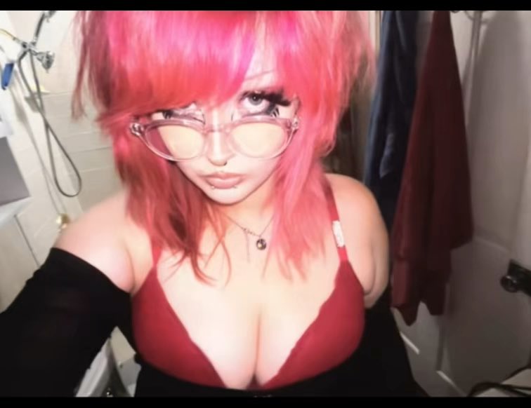 A person with bright pink hair, wearing clear-framed glasses and a low-cut maroon top, takes a selfie in what appears to be a bathroom. The image relates to the post as a promotional photo for a "moothunt" account, with hashtags like #promotwt, #gamingtwt, and #animetwt indicating the account's focus on gaming and anime content.