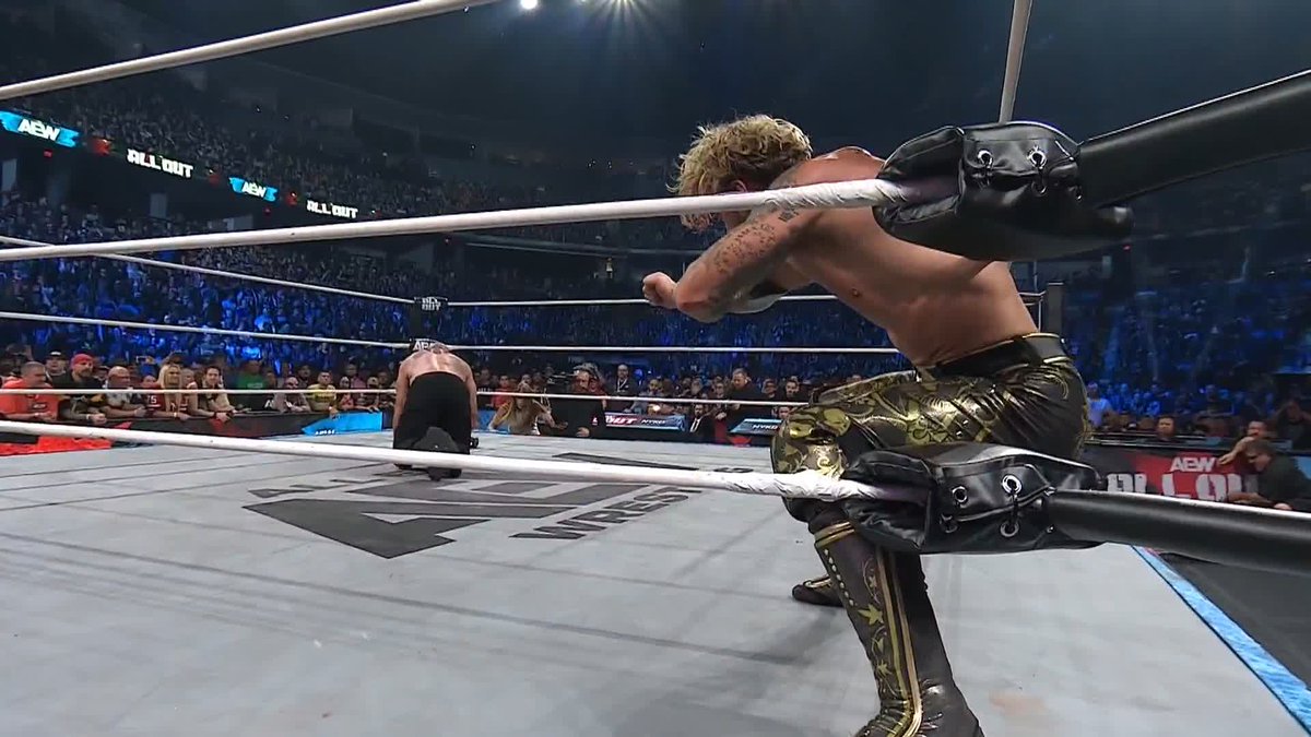 A blonde wrestler in ornate gold pants clutches the ring ropes during an AEW All Out match, while his opponent kneels on the mat below, with "AEW All Out" signage visible around the arena.