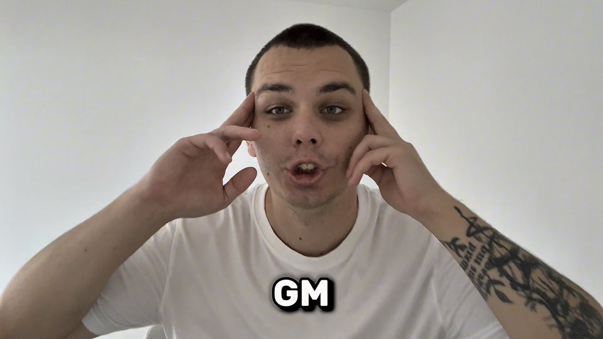 A young man with a short haircut and tattooed arm pointing to his temples with both hands, wearing a white t-shirt, with 'GM' text overlaid.