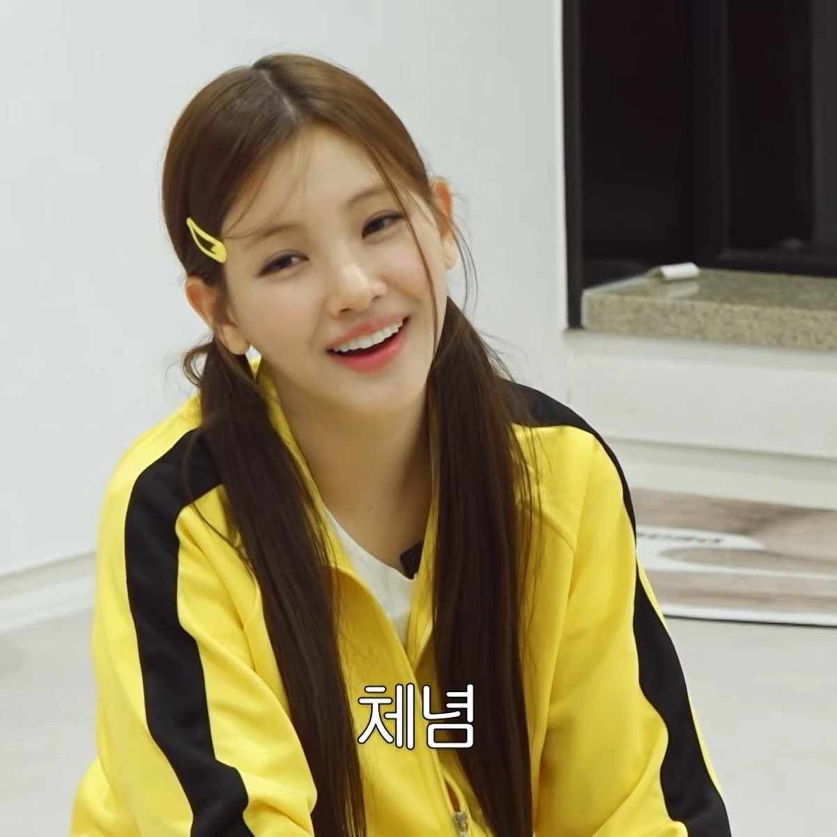 A smiling woman with long brown hair wearing a yellow track jacket with black stripes and a yellow hair clip, with Korean text '체념' displayed.