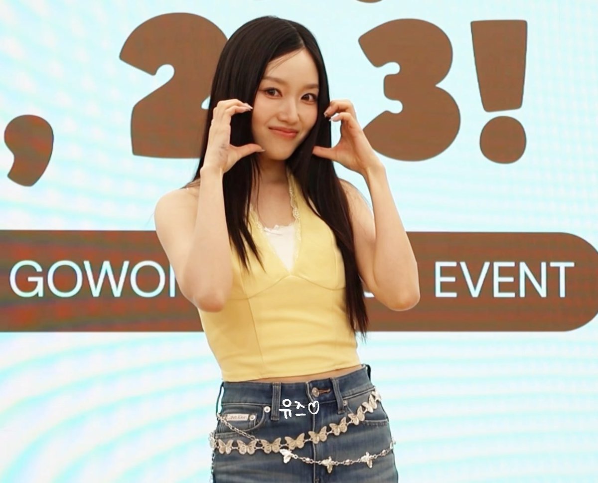 A young woman with long dark hair posing with her hands forming a heart shape at what appears to be a GOWON event, wearing a yellow halter top and denim jeans with decorative chain.