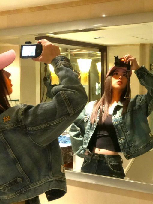 A mirror selfie of a young woman wearing a denim jacket and cap, taking the photo with a digital camera, with another person in a pink cap visible on the left.