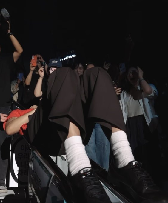 A low-angle shot of a person in a dark skirt and white knee-high socks sitting at the edge of a stage, surrounded by fans taking photos with their phones.