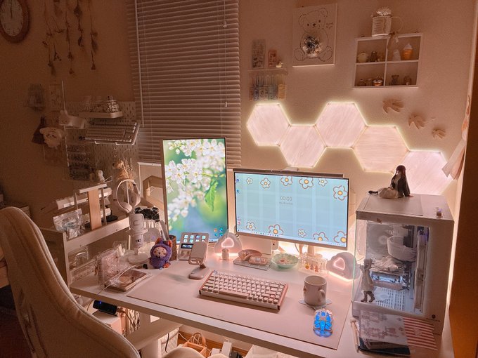 An aesthetic pink-themed desk setup with two monitors displaying cherry blossom and flower wallpapers, a white mechanical keyboard, hexagonal LED wall lights, and various cute figurines.
