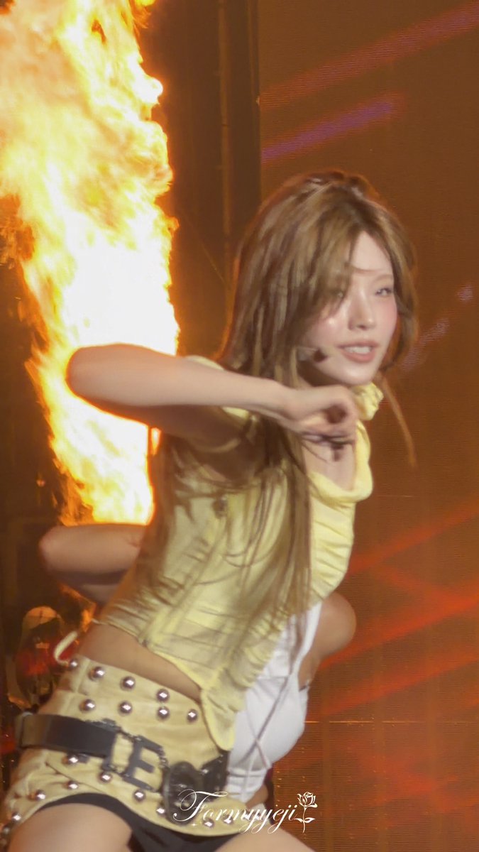 A female performer in a yellow outfit dances on stage with a pyrotechnic flame shooting up behind her; the image is watermarked "Teruyagi".