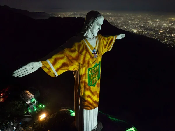 The iconic Christ the Redeemer statue in Rio de Janeiro is illuminated at night, wearing a golden football jersey featuring the NFL shield logo and "A Equipe Tudo NFL" text across the front, with the sprawling city lights of Rio visible below. The image directly illustrates the post's reference to the NFL game in Rio, showing the famous monument dressed in team colors to honor the American football event being held in the city.