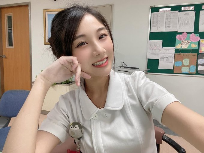 A young woman in a white nurse-style uniform smiles at the camera while making a phone gesture with her hand, in what appears to be a medical or office setting with a bulletin board behind her.